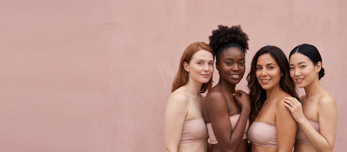 Four smiling women of different skin tones standing side by side against a pink wall
