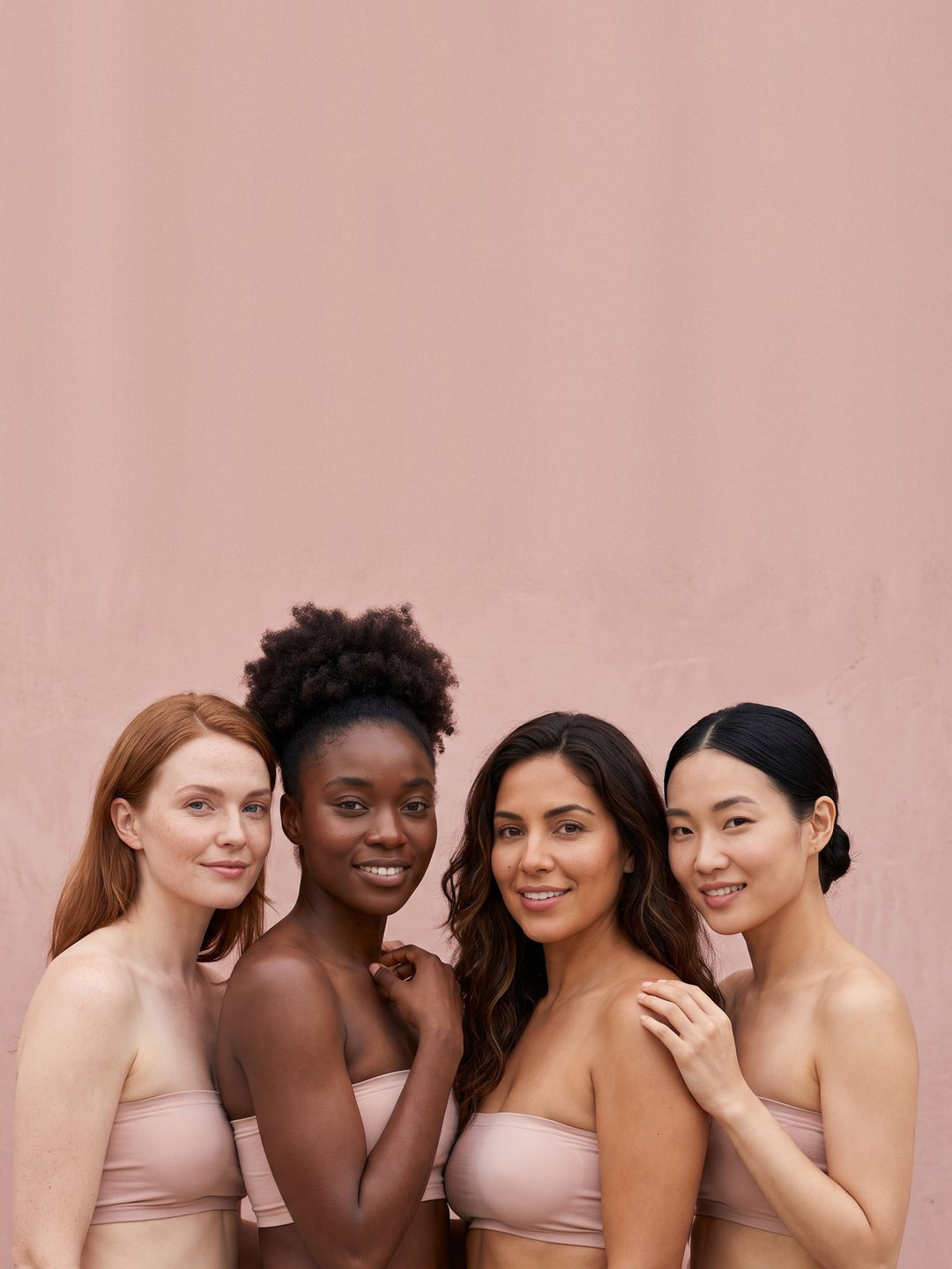 Four smiling women of different skin tones standing side by side against a pink wall